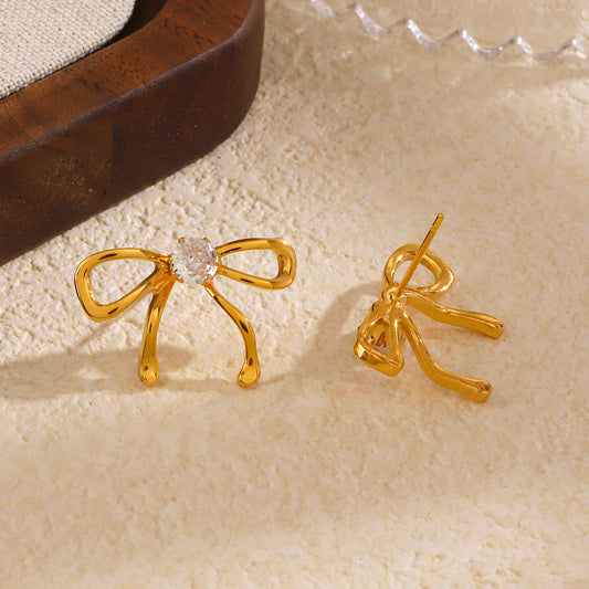 Gold bow-shaped earrings with a central gemstone on a beige surface.