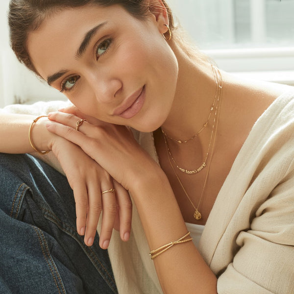 Woman wearing gold jewelry including necklaces, bracelets, and rings, with a blurred indoor background.