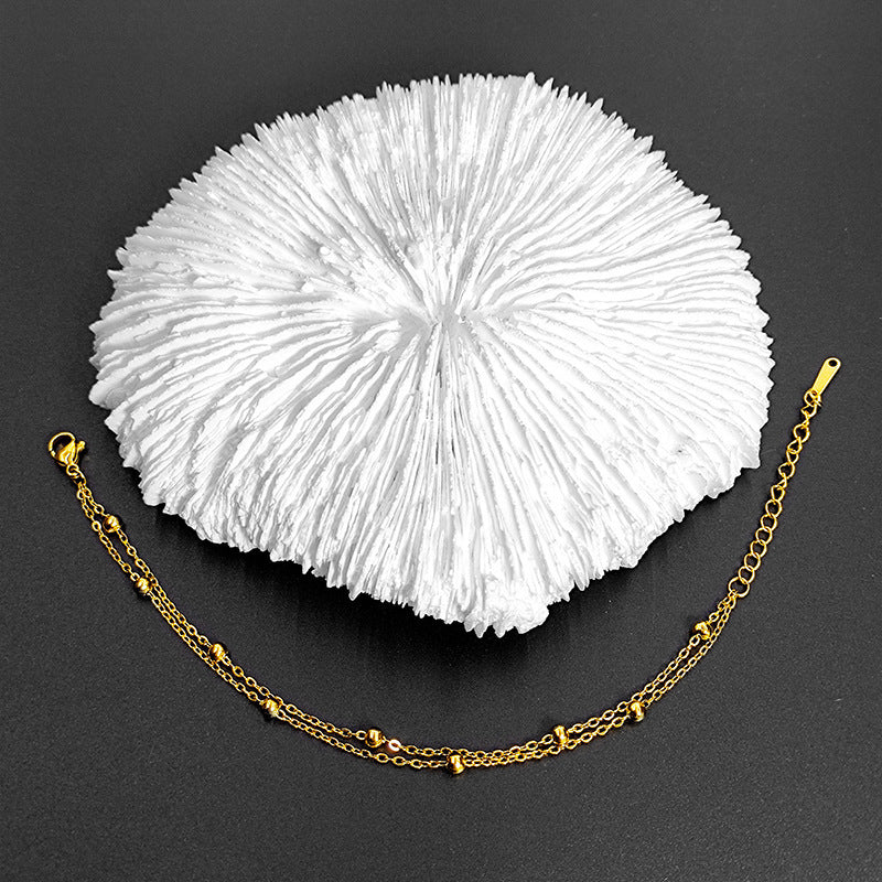 Gold double-chain bracelet with beads, styled on white coral against a dark, textured background